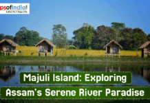 Majuli Island: Exploring Assam’s Serene River Paradise Scenic view of Majuli Island in Assam featuring traditional wooden huts on stilts beside a calm water body, surrounded by lush green fields and trees under a clear blue sky.