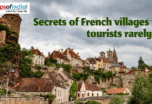 Secrets of French Villages That Tourists Rarely See Hillside view of a hidden French village with stone tower, colorful houses, church spires, and a bridge under a cloudy sky.