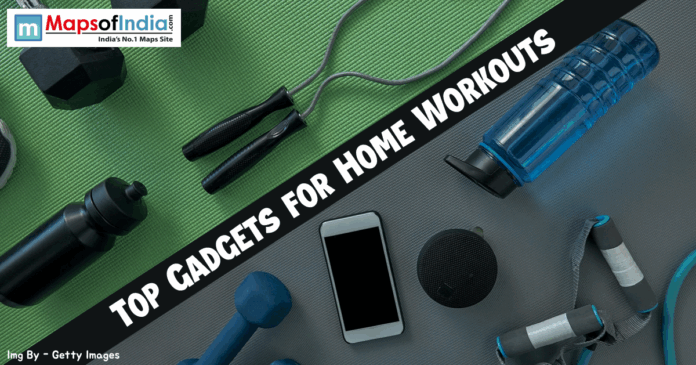 Top Gadgets For Home workout Top-down view of fitness equipment including dumbbells, a jump rope, resistance bands, a smartphone, and water bottles on a green and grey gym mat with the text "TOP GADGETS FOR HOME WORKOUTS."