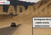 The Magnetic Hill of Ladakh: Gravity Defied or Illusion Car driving on a desert highway in Ladakh near Magnetic Hill with mountains in the background and a sign reading “The Magnetic Hill of Ladakh: Gravity Defied or Illusion.”